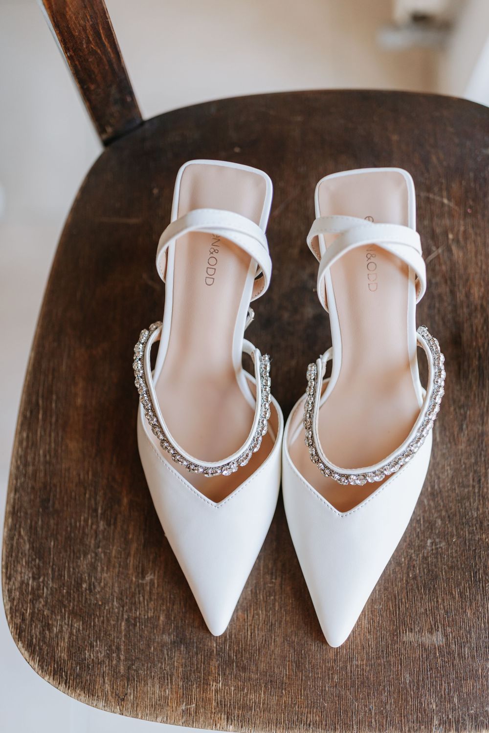White pointed-toe bridal shoes with crystal embellished ankle straps displayed on rustic wooden surface.