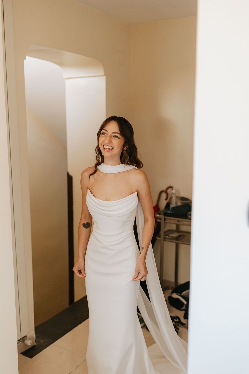 A person wearing a white wedding dress stands in a light-filled room with mirrors, showing multiple angles.