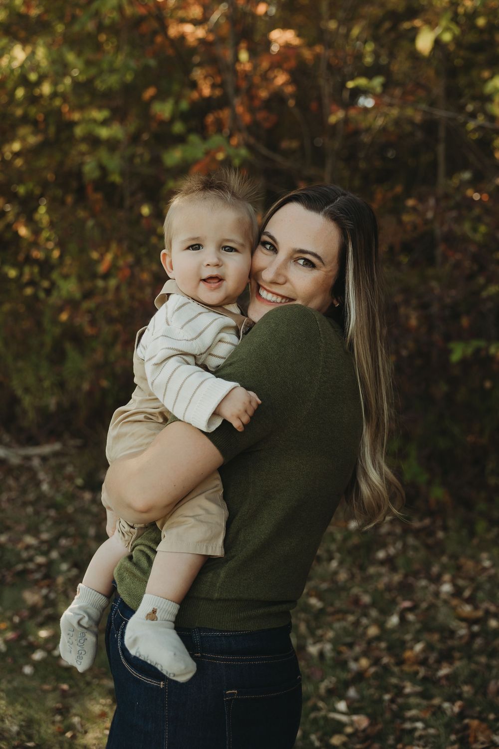 A parent in a dark green sweater holds a baby wearing light colored clothing against an autumn forest backdrop.