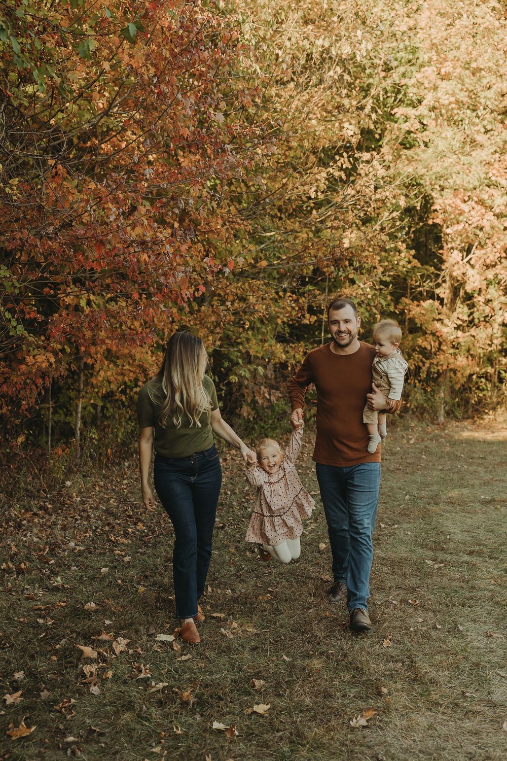 A family walks together on an autumn path surrounded by colorful fall foliage and golden sunlight.