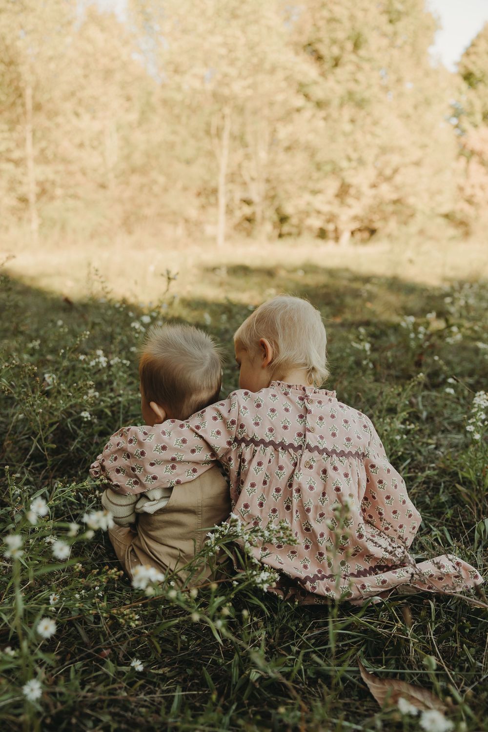 Two young children lie together in green grass wearing vintage-style clothing in a dreamy outdoor setting.