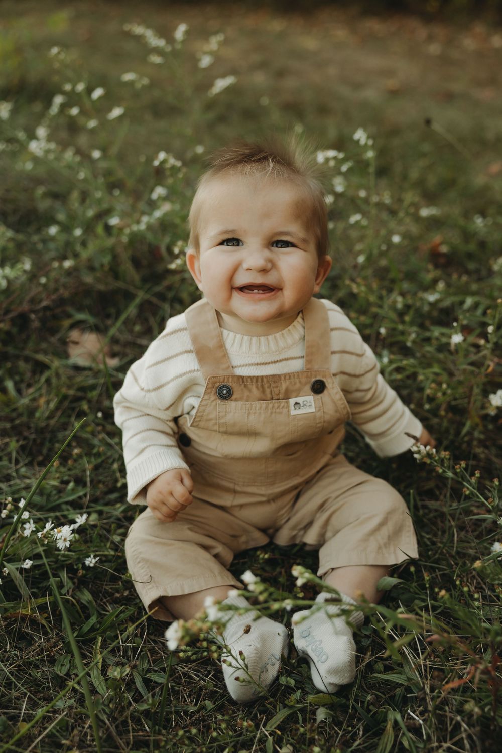 A small child in tan overalls and white sneakers sits on grass with white flowers while smiling at the camera.