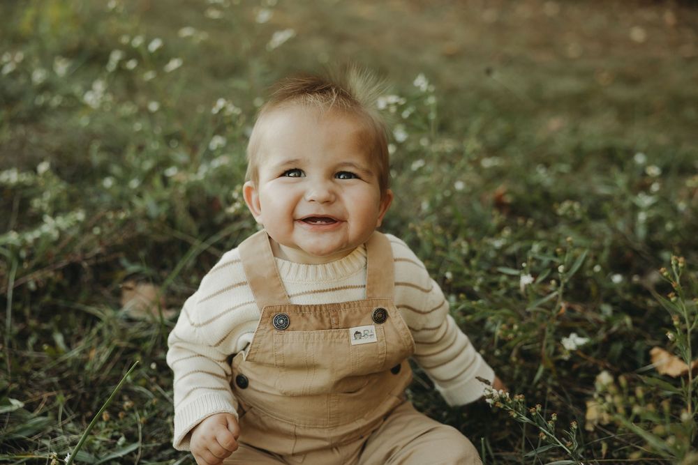 A baby wearing tan overalls sits on grass during an autumn outdoor photo session.