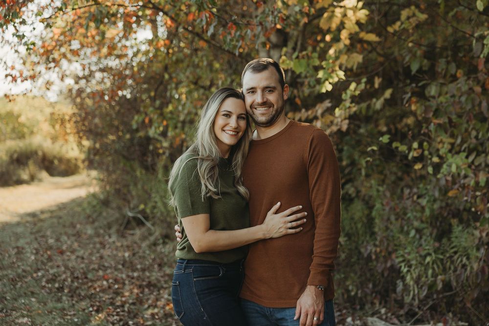 A couple embraces on an autumn path with golden sunlight filtering through trees in the background.