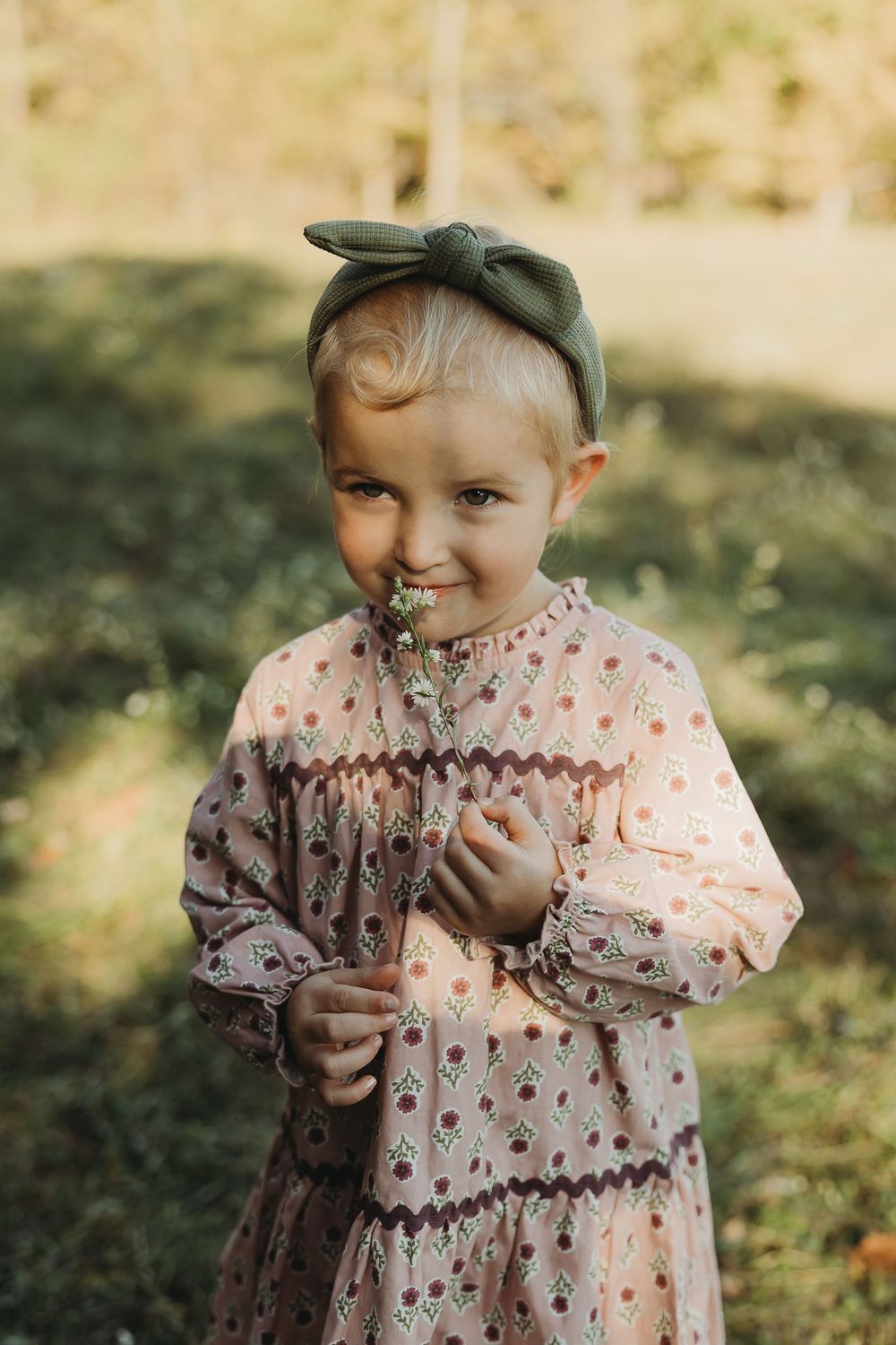 A young child in a patterned dress and bow headband holds wildflowers while standing in dappled sunlight outdoors.
