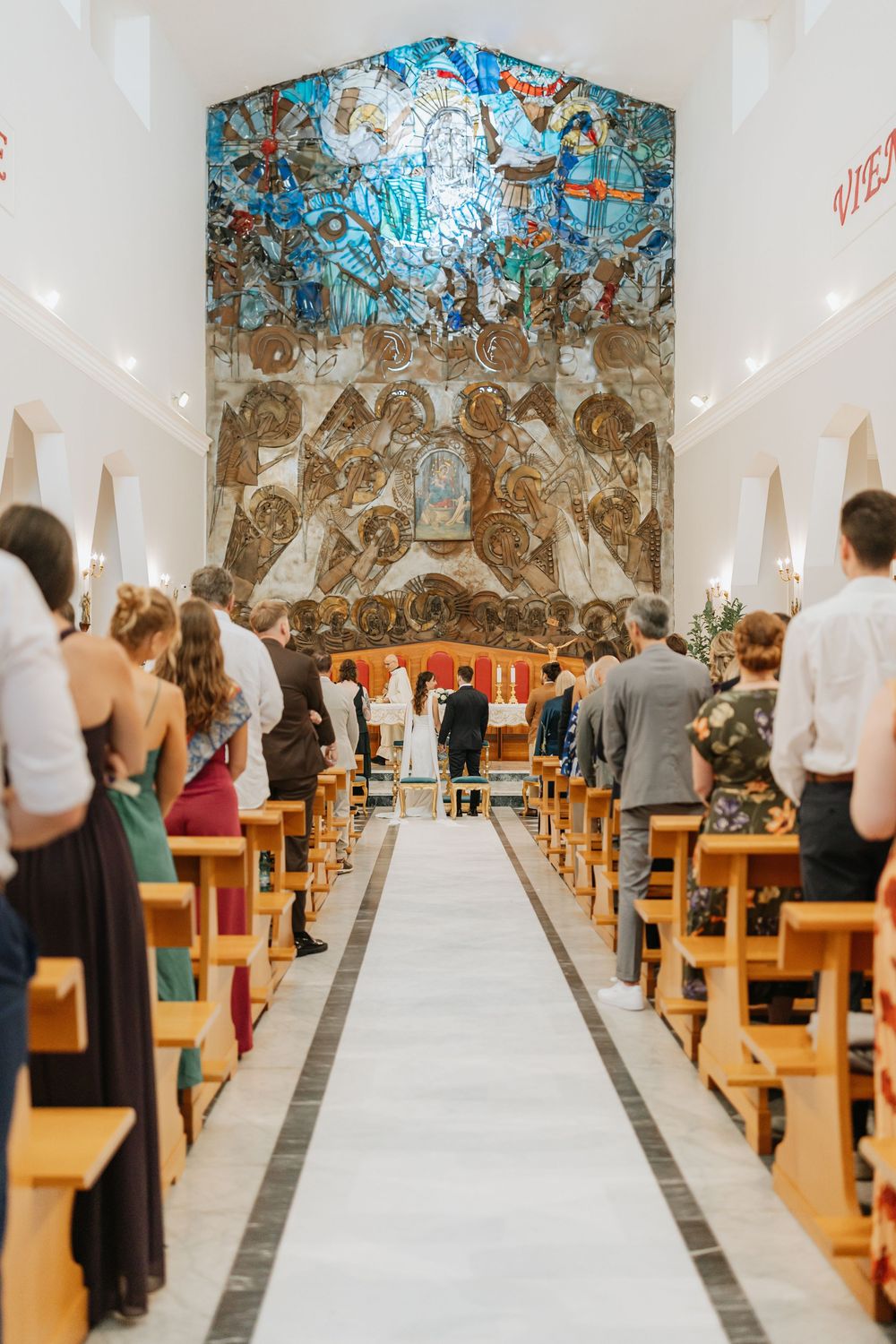 Panoramic view of a modern church interior with white walls, wooden pews, and decorative ceiling artwork during a service.