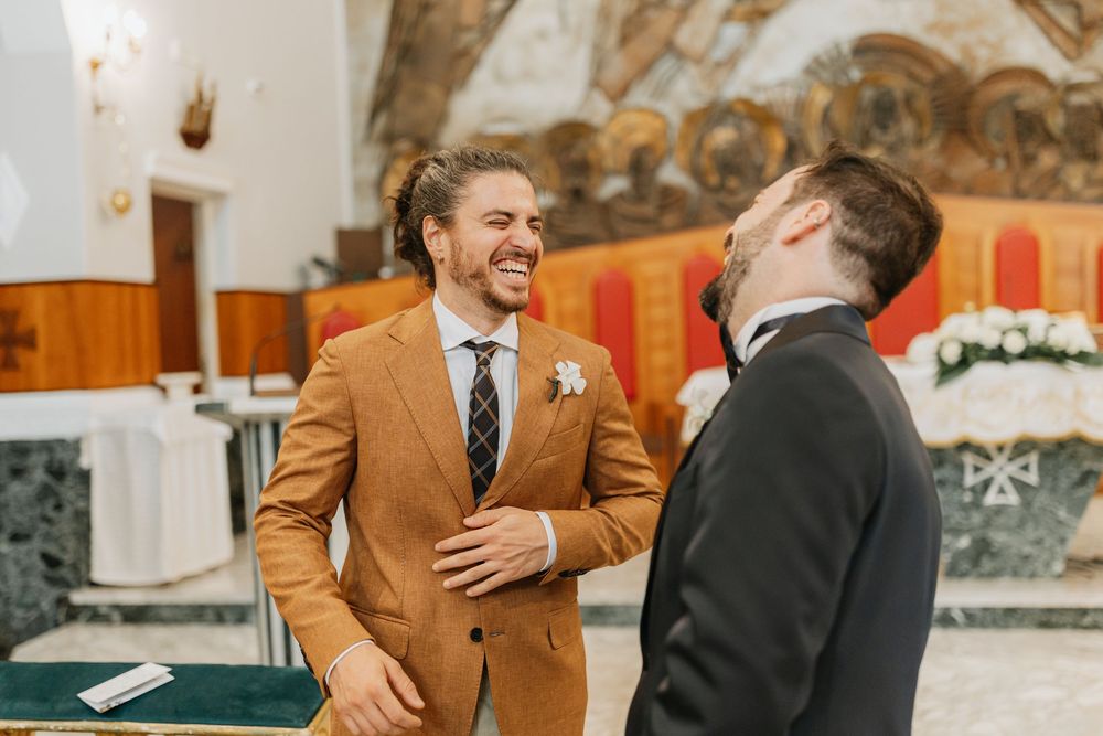 Groomsmen in black suits and one in brown suit laugh and embrace during a wedding celebration.