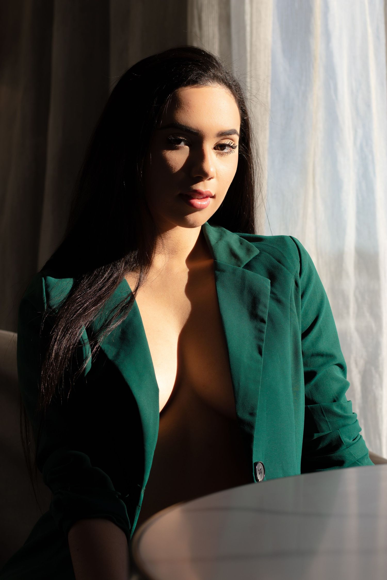 Woman in emerald green blazer poses by window in dramatic lighting.