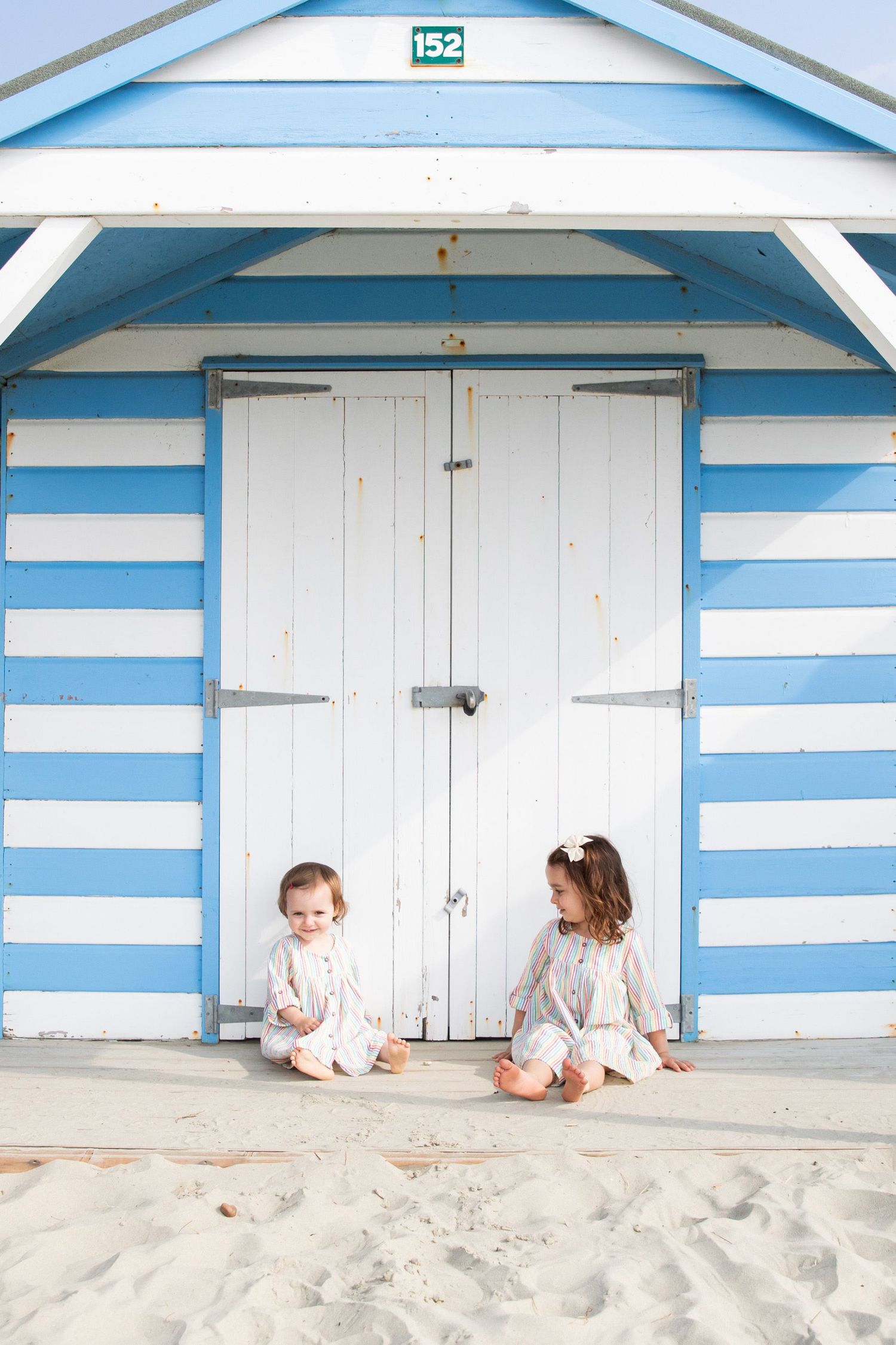 Children sitting in front of a blue and white striped beach hut door on sandy ground.