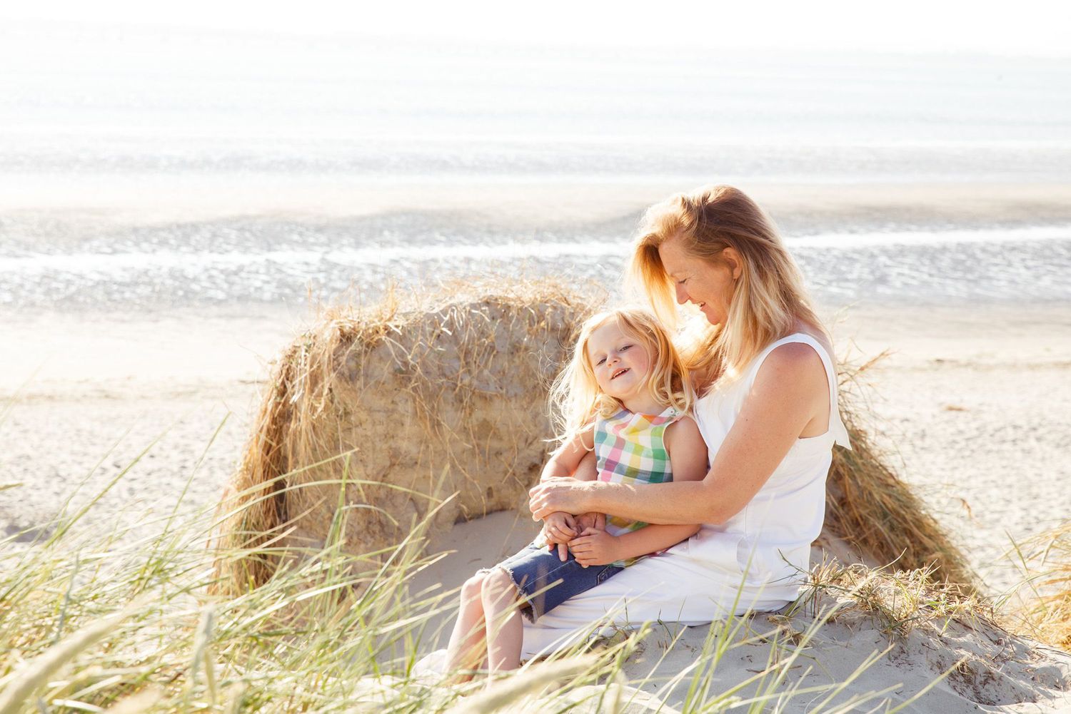 Two people cuddling on beach dunes surrounded by coastal grass at sunset.