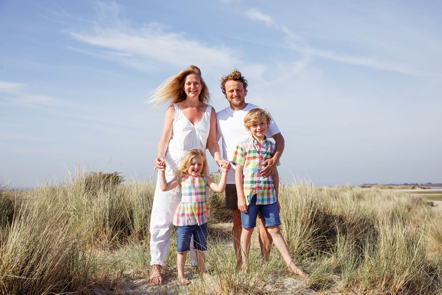 Family of four standing together among beach grass dunes against blue sky.