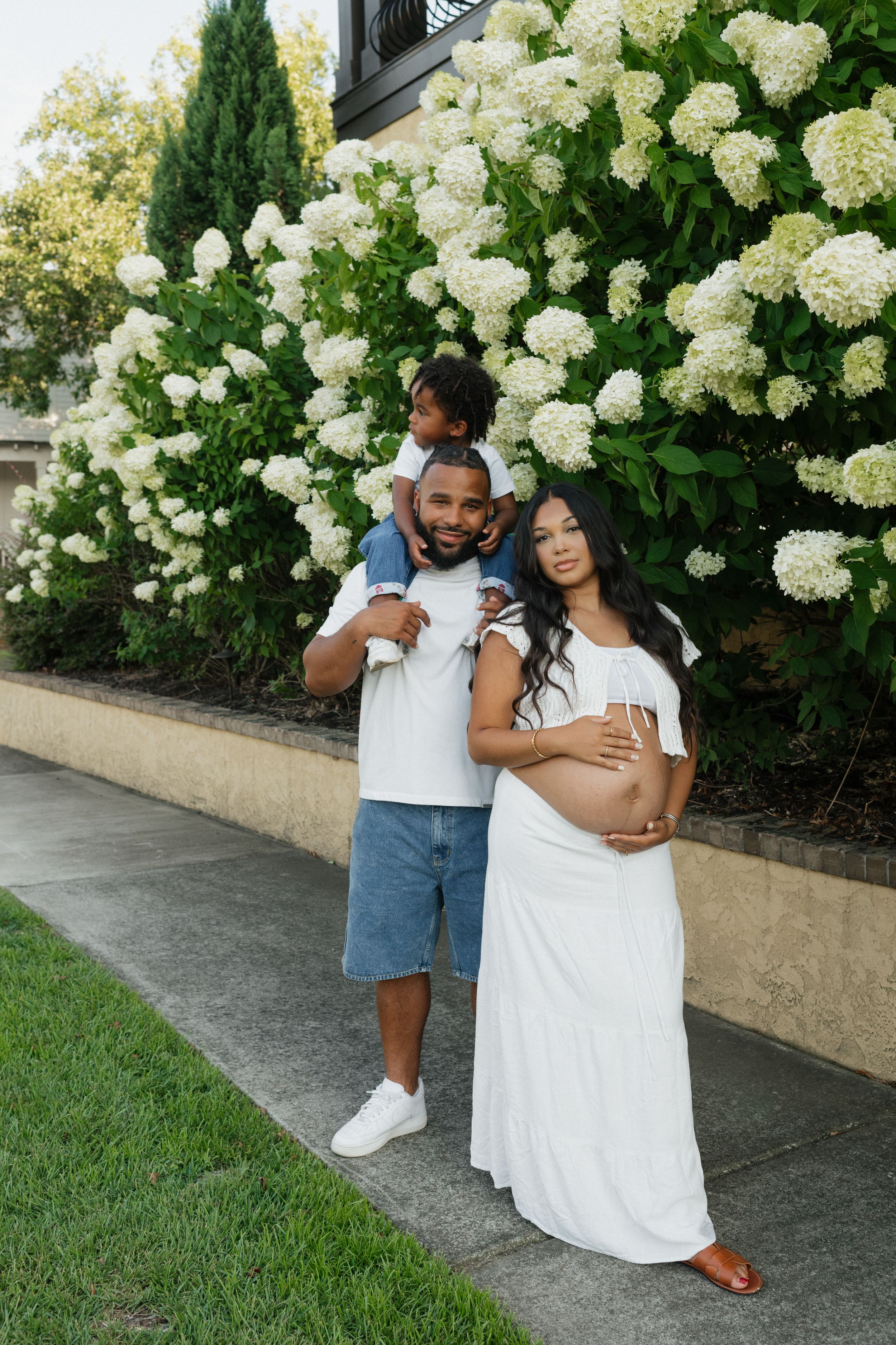 Couple posing by blooming white hydrangea bushes in casual summer attire.