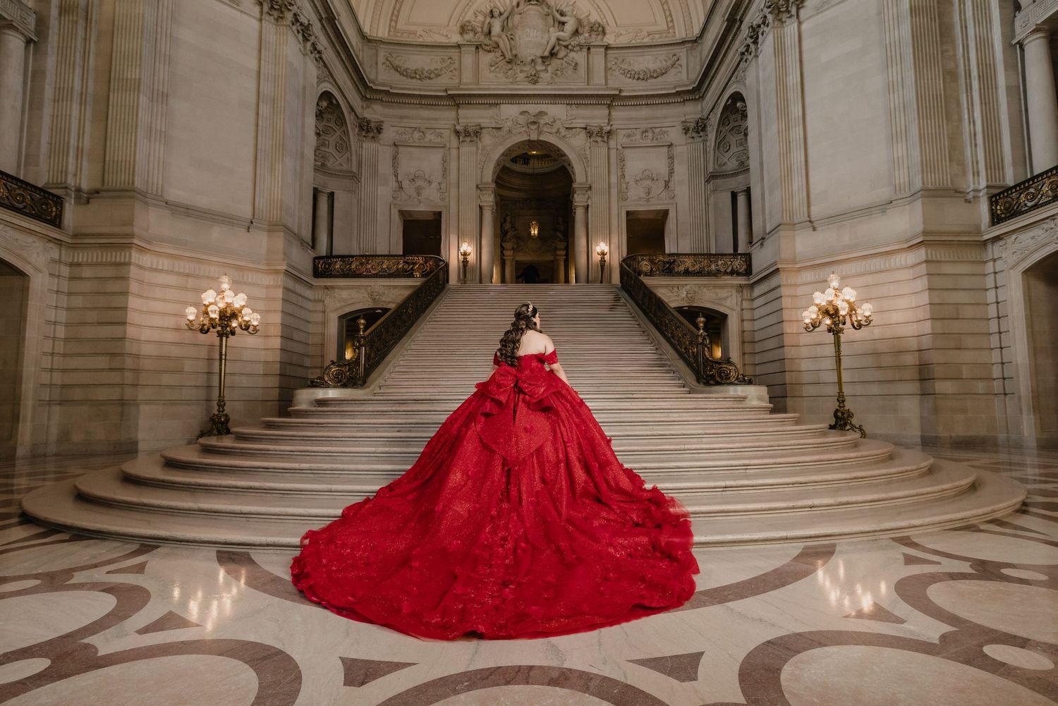 A stunning red ballgown with a dramatic train flows across the marble staircase of an elegant historic building interior.
