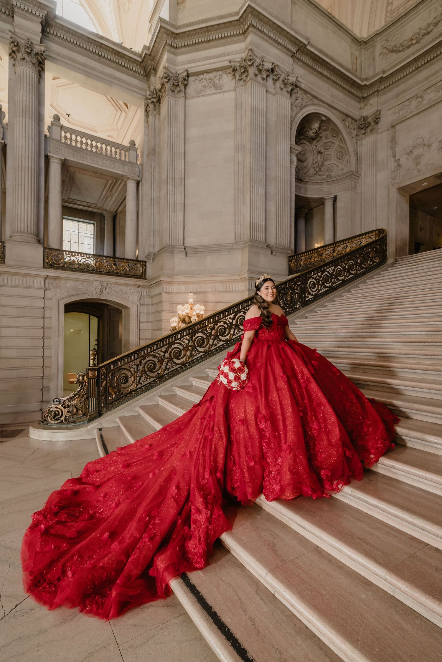 A flowing red ballgown with a dramatic train cascades down elegant marble stairs in a grand architectural setting.