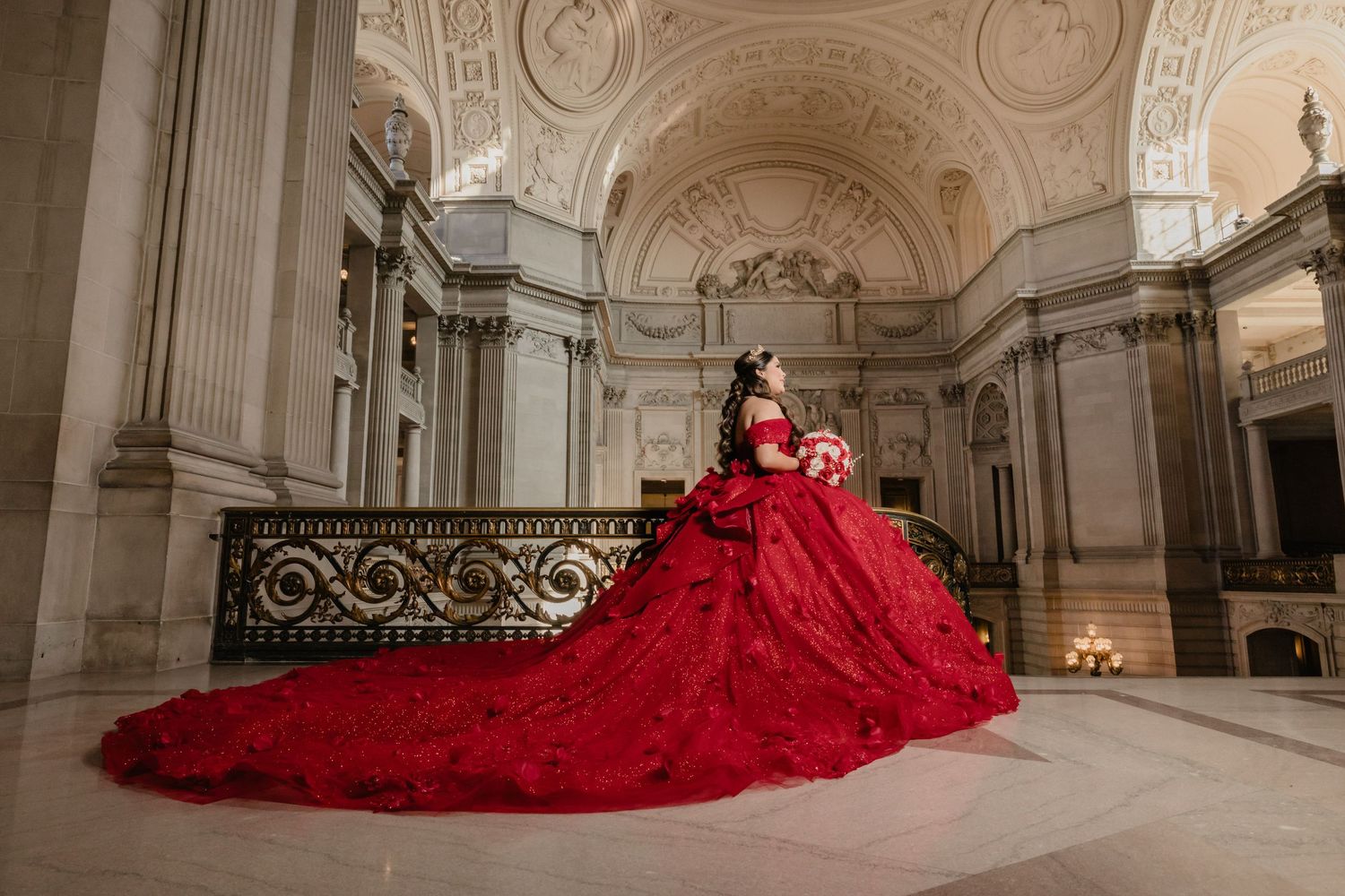 A stunning red ballgown dress with a dramatic train flows across an ornate marble floor in a grand historic building.