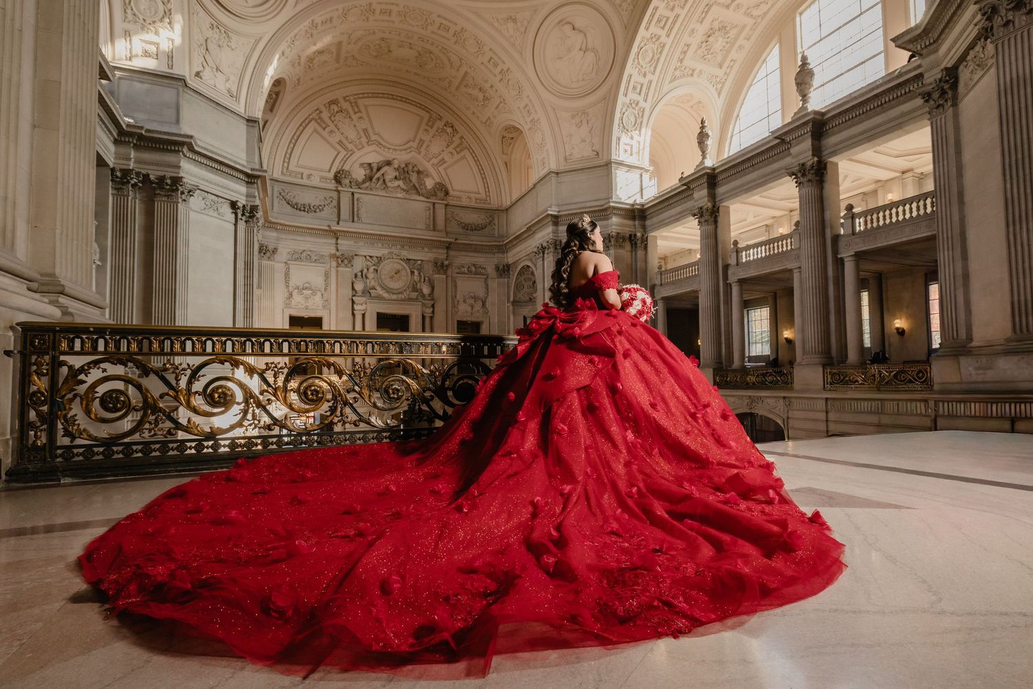 A stunning red gown with an expansive train flows across a grand marble hallway in an ornate classical building.