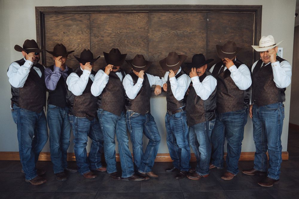 Row of groomsmen in cowboy boots and jeans lined up against rustic wall.