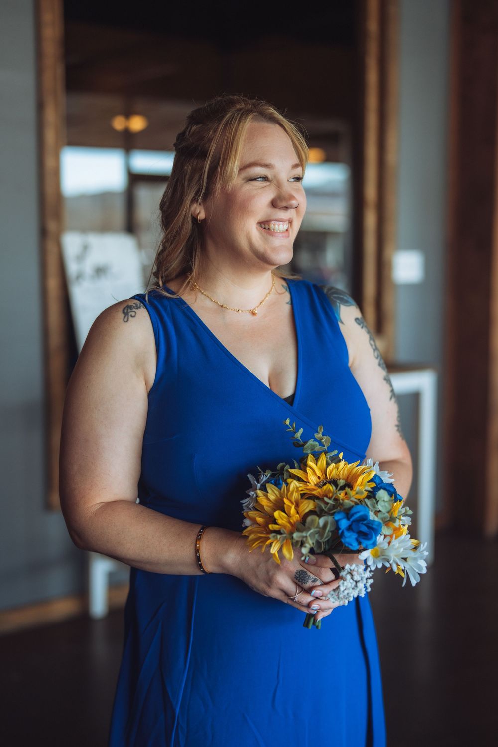 Bridesmaid in sleeveless blue dress carrying sunflower and white daisy bouquet.