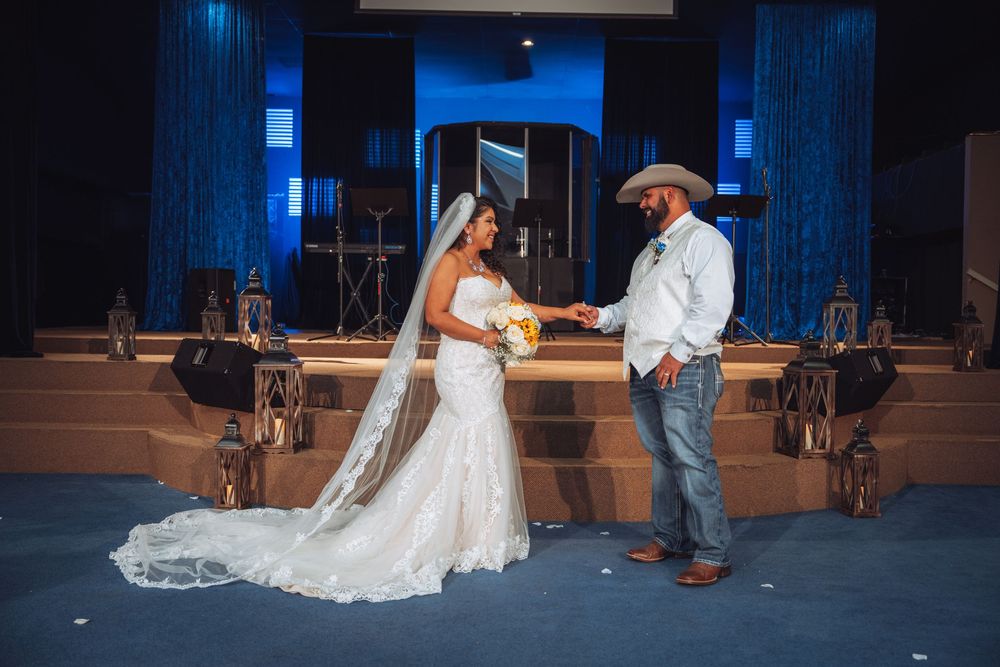 A bride and groom hold hands during their wedding ceremony on a blue-lit stage.