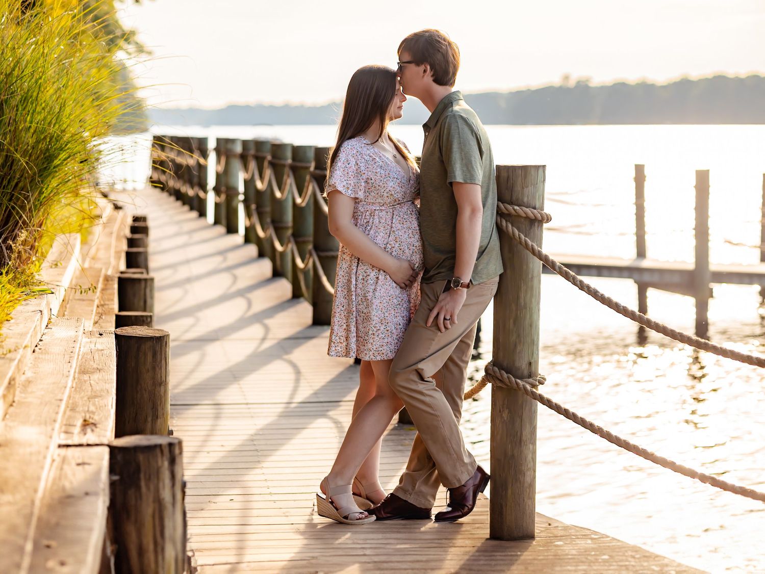 Couple in light summer attire embraces on a wooden dock at sunset with golden sunlight streaming through trees.