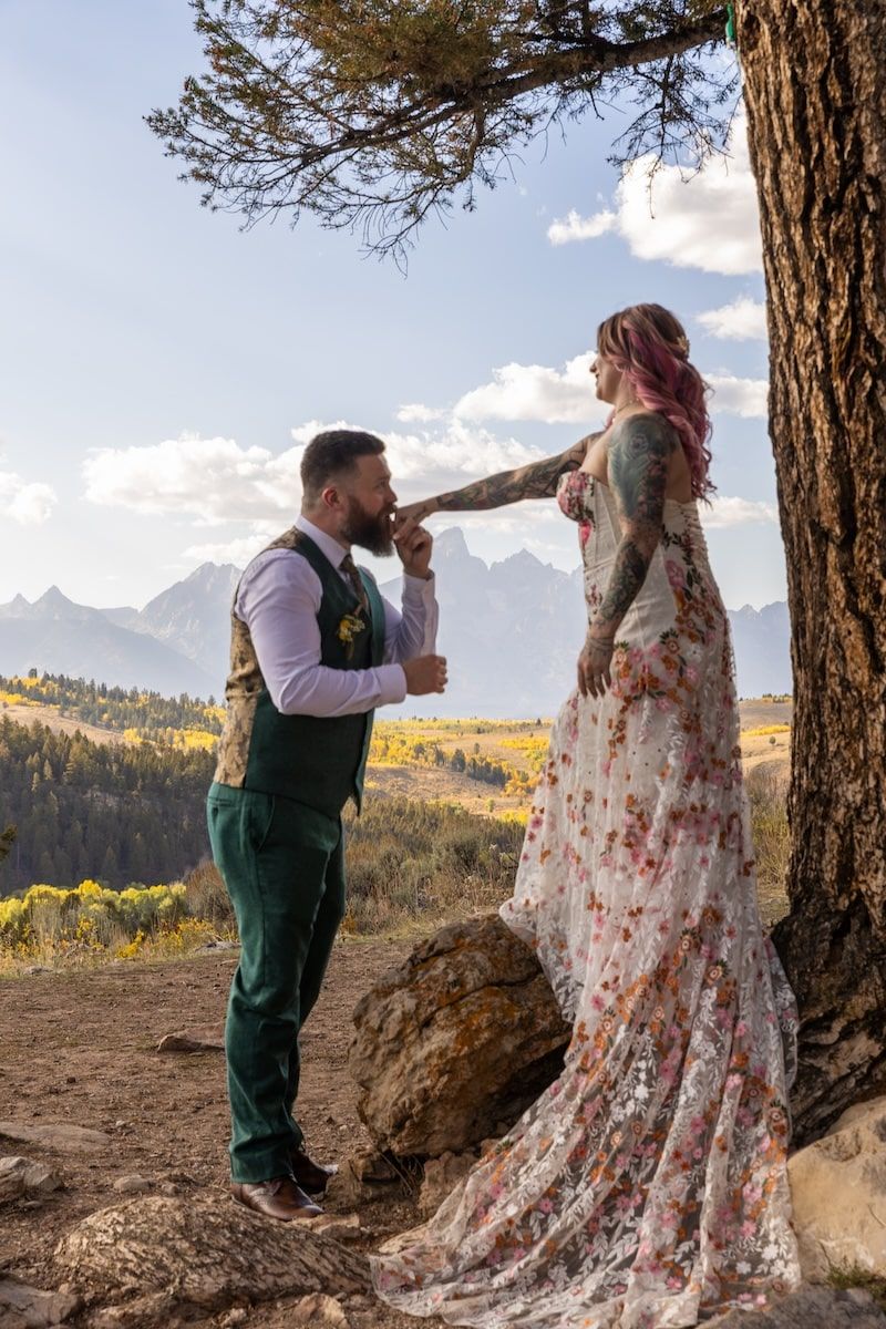A couple shares a romantic moment in front of a pine tree with scenic mountain views in the background.