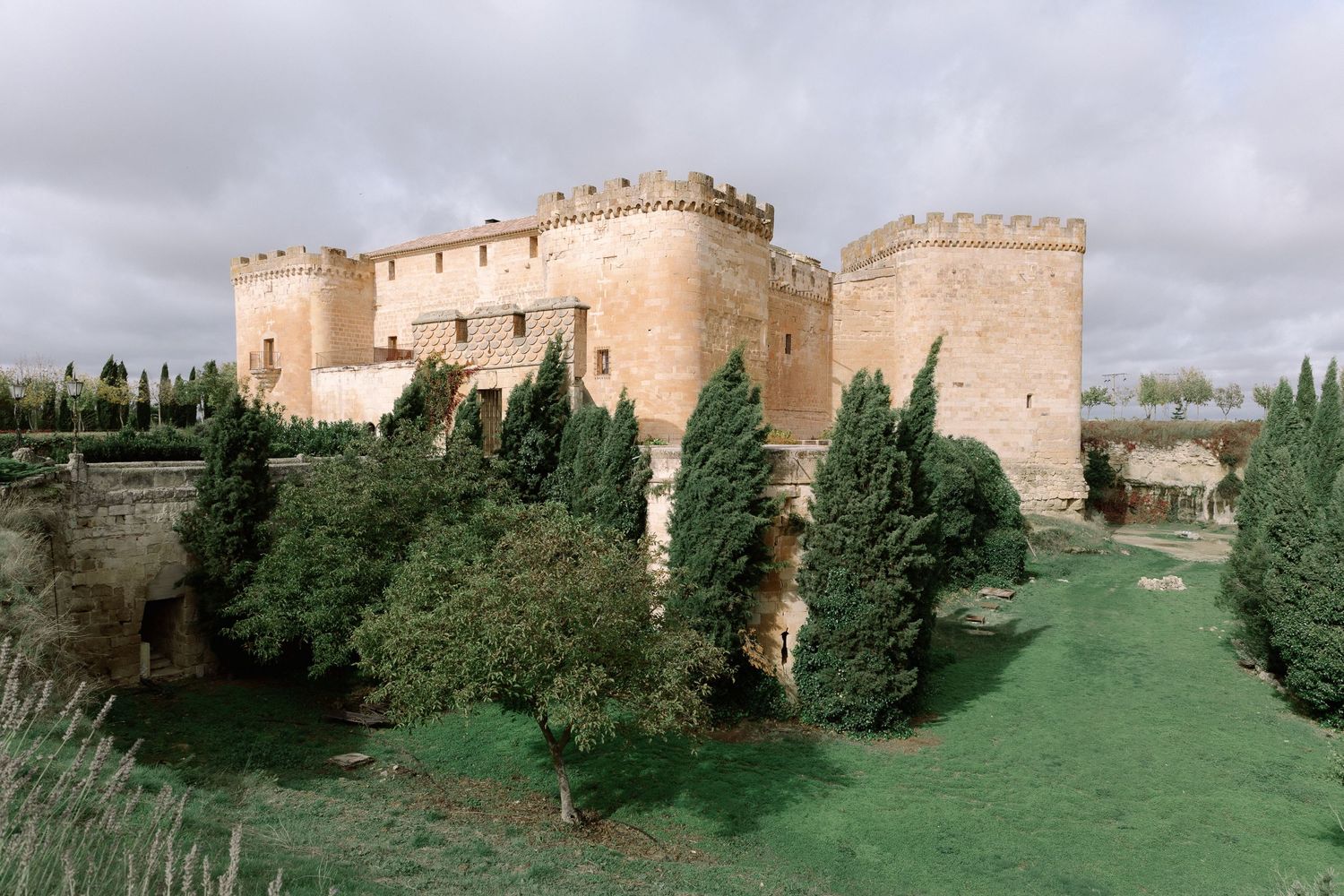 Fortified medieval castle with stone towers and turrets surrounded by cypress trees and green lawns in Spain.