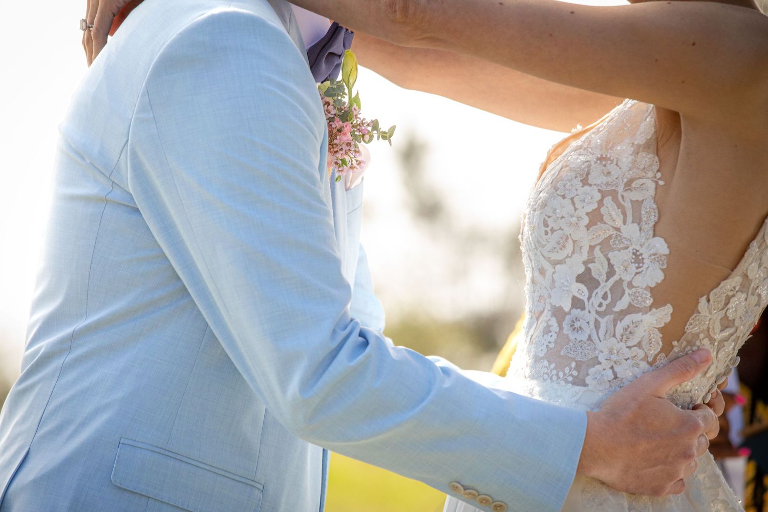 Bride and groom sharing a romantic embrace, wearing lace wedding dress and light blue suit outdoors.