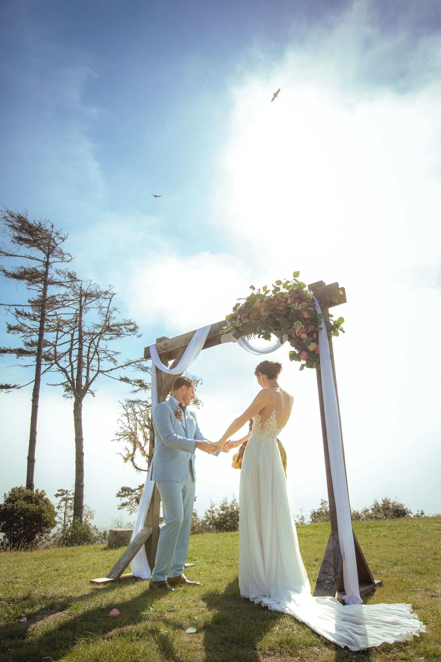 Couple holding hands under a floral arch during an outdoor wedding ceremony on a sunny day.