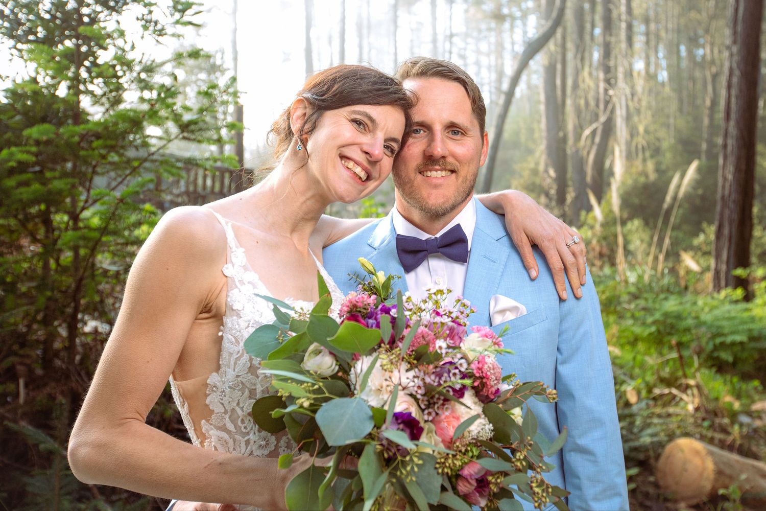 Happy bride and groom pose together outdoors in forest setting, bride holding colorful bouquet, groom in light blue suit.