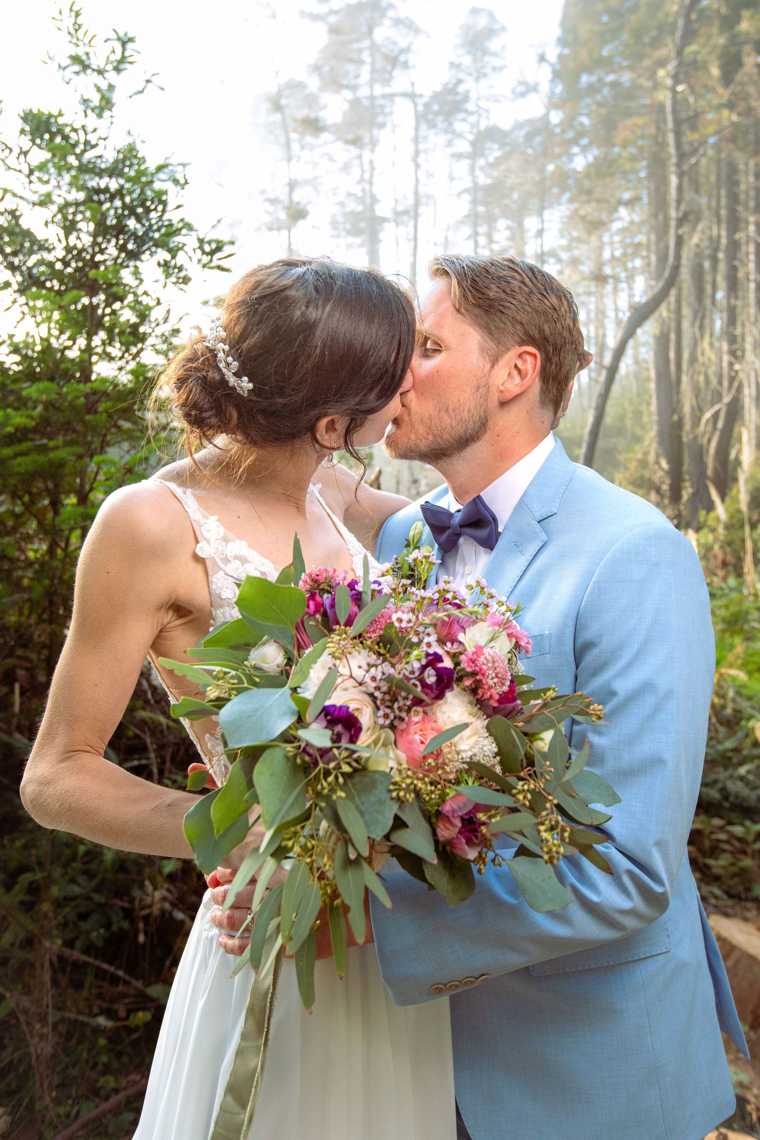 Bride and groom sharing a kiss outdoors, with bride holding a colorful bouquet of flowers in a forest setting.