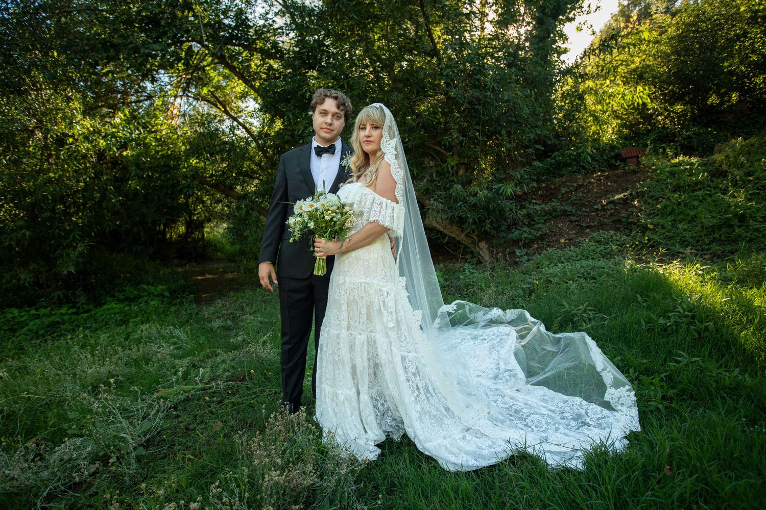Newlyweds posing on grassy hillside with flowing white veil in afternoon light.