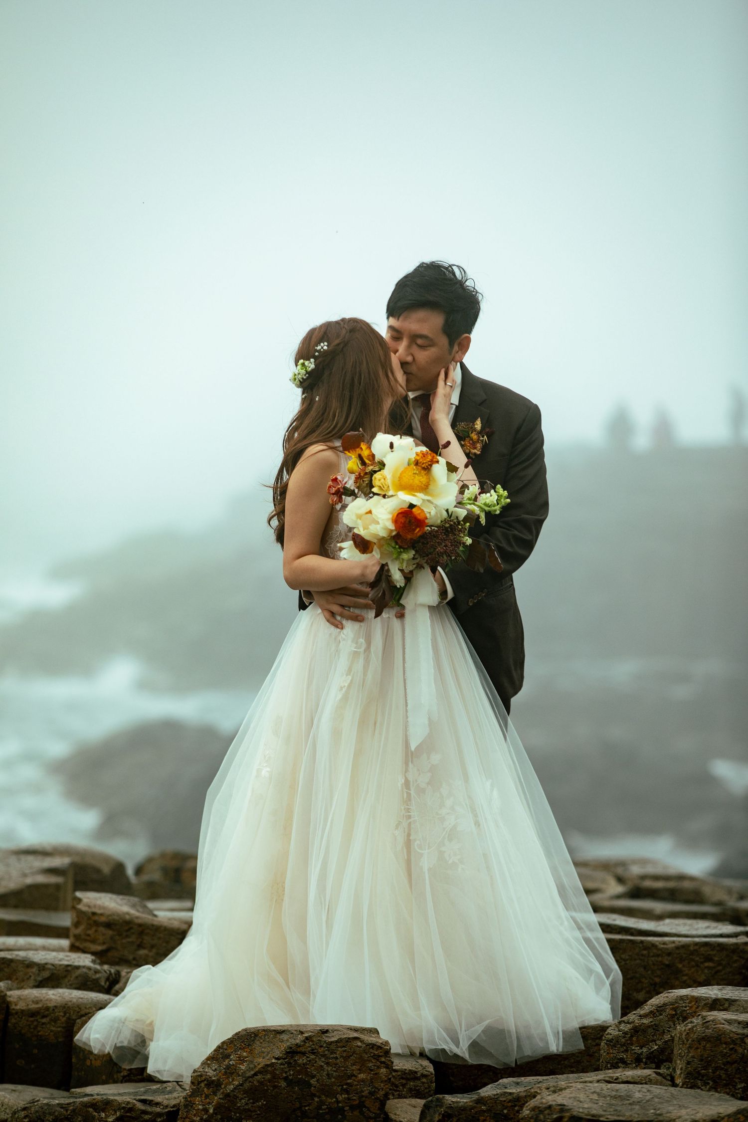Romantic coastal wedding portrait with misty cliffs and ocean backdrop.