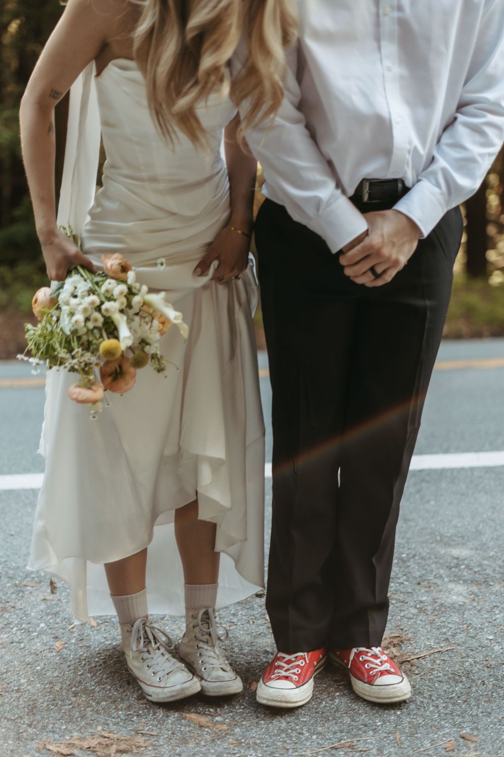 A couple in wedding attire wearing matching Converse sneakers stands together holding a white floral bouquet.