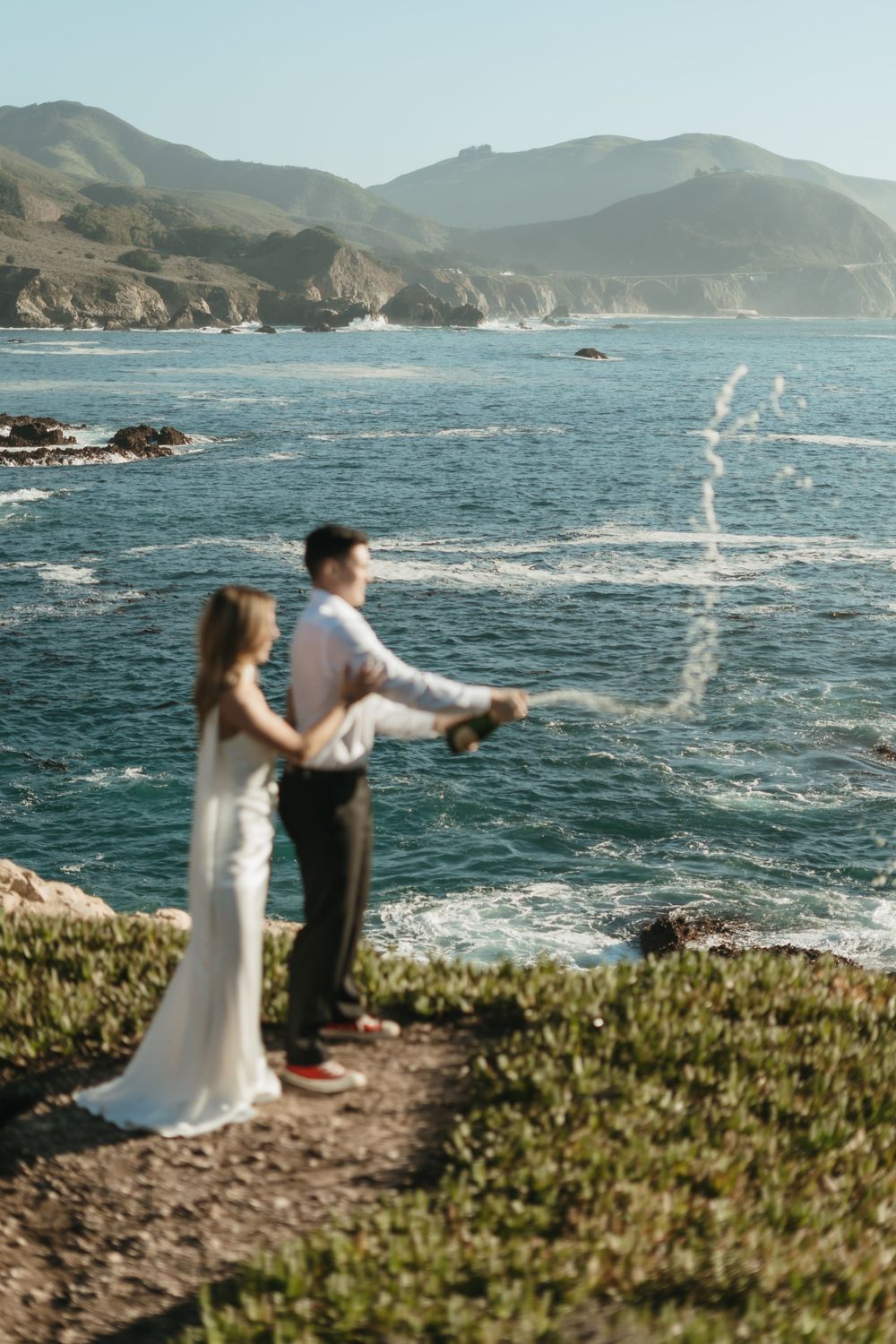 A couple stands at the edge of a coastal cliff overlooking dramatic ocean waves and rocky coastline.