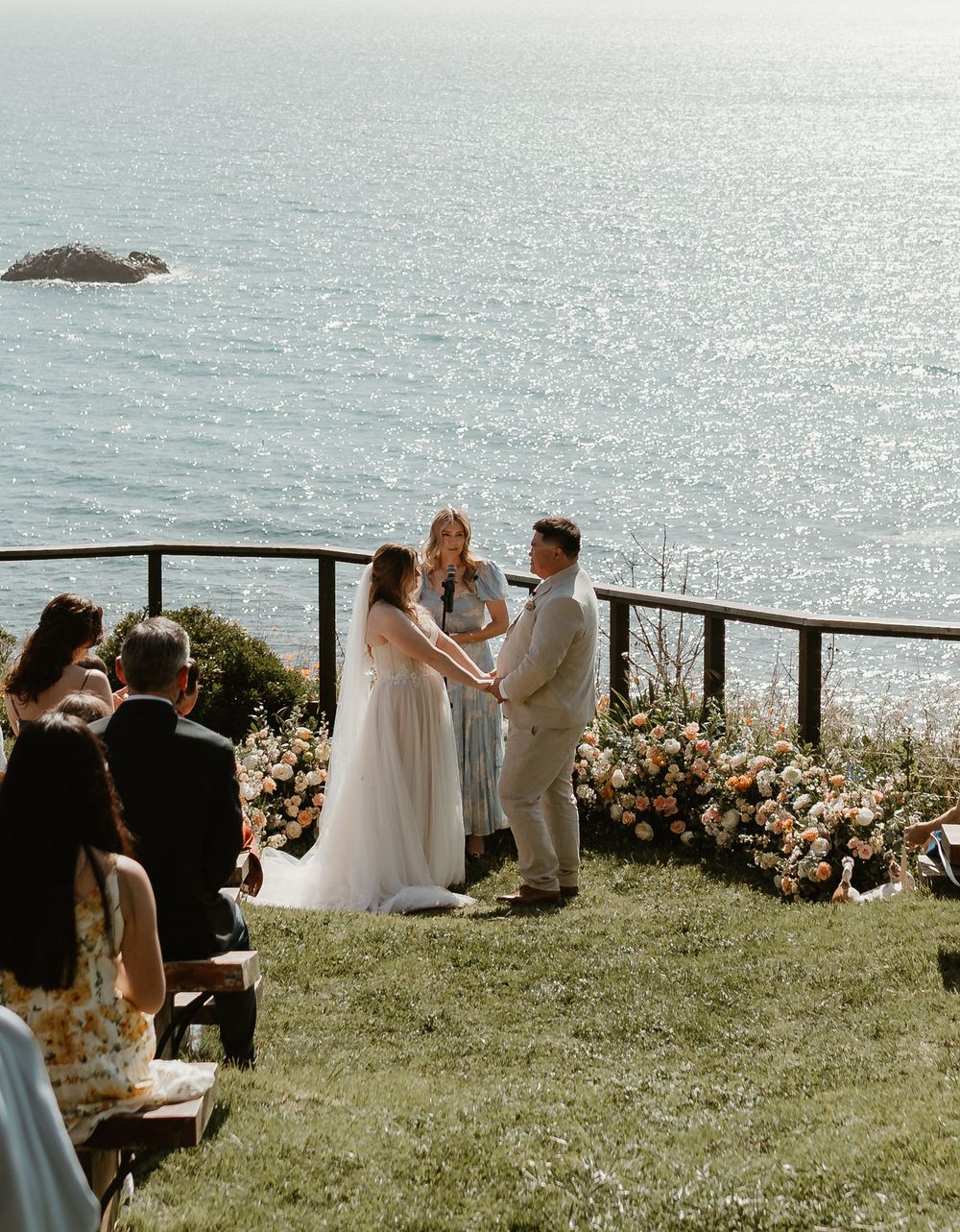 A coastal wedding ceremony takes place on a scenic cliff overlooking the sparkling ocean on a sunny afternoon.