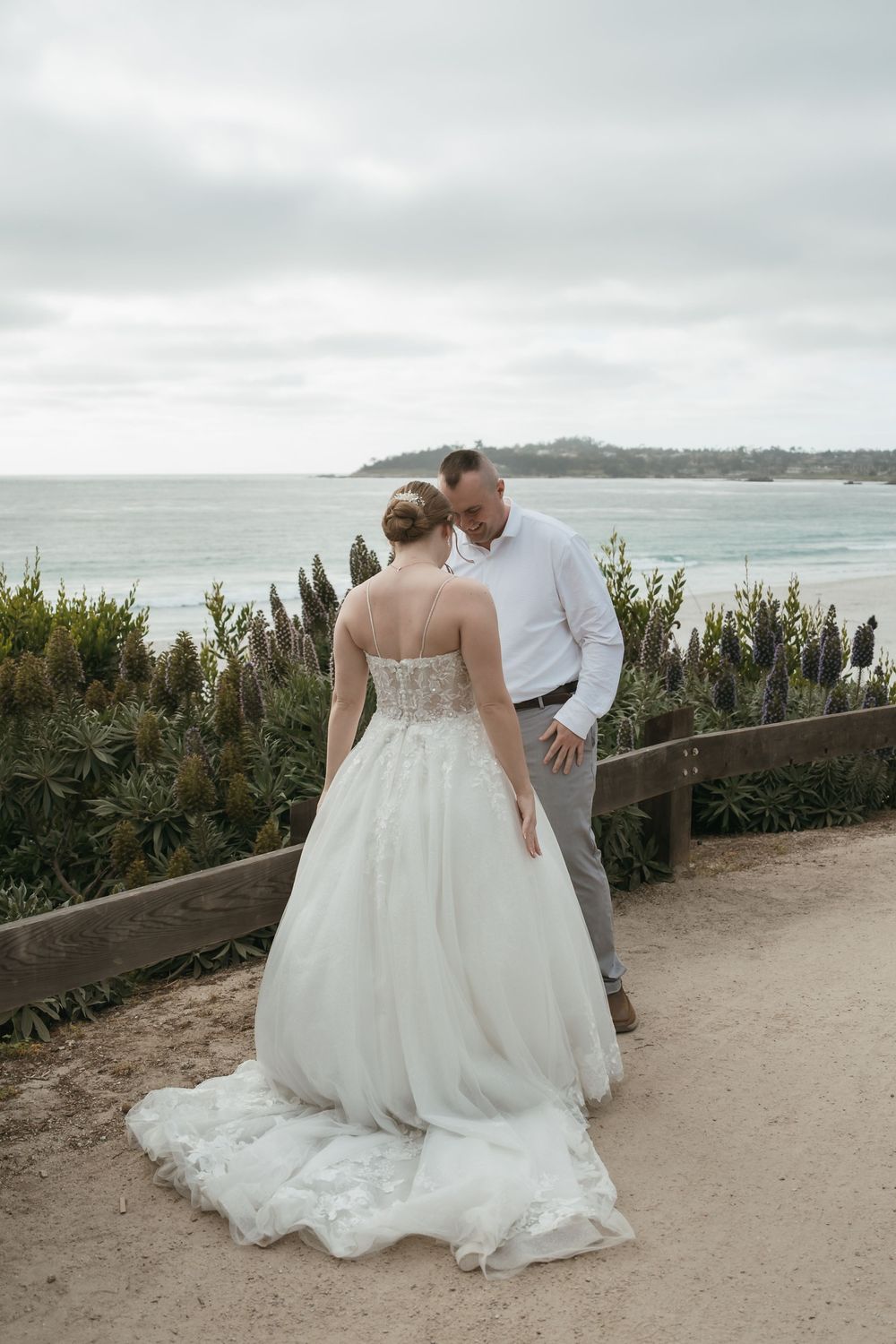 Bride in white gown and groom in white shirt share a romantic moment on a coastal path with ocean views.