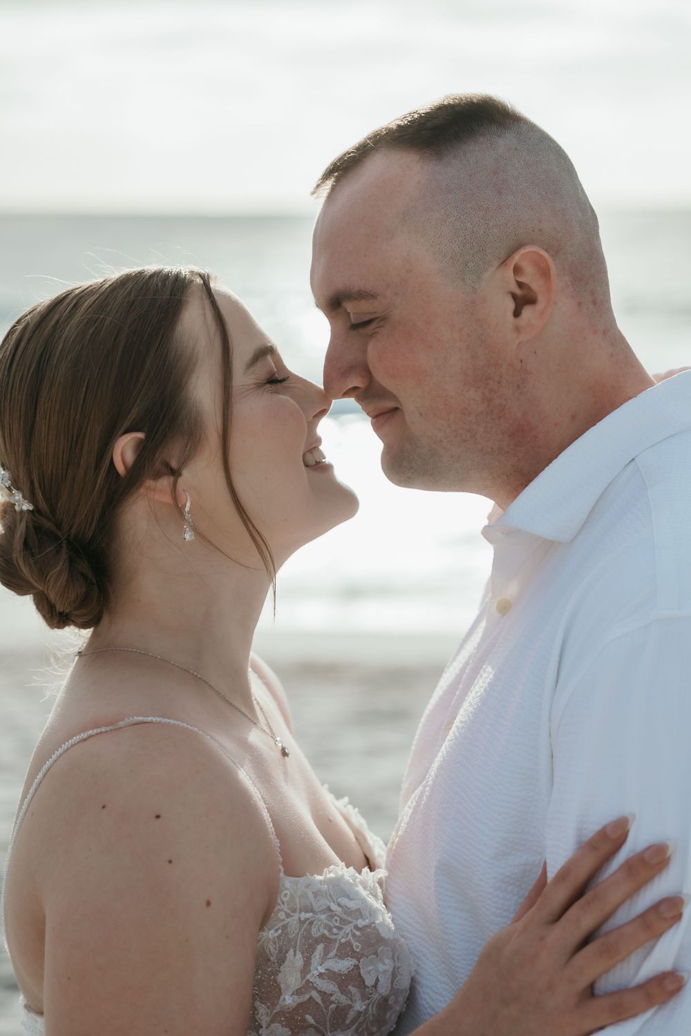 A couple shares a romantic almost-kiss on a beach, the bride in lace and groom in white, with ocean waves behind them.