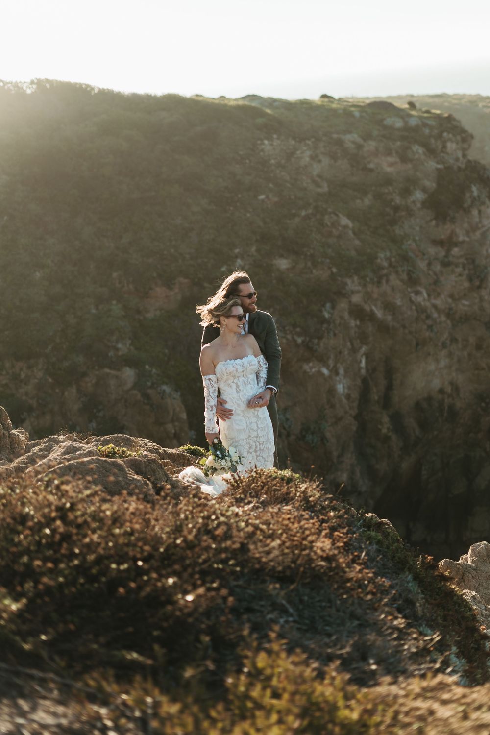 A person in a white dress stands atop a scenic mountain ridge during golden hour, photographing the landscape.