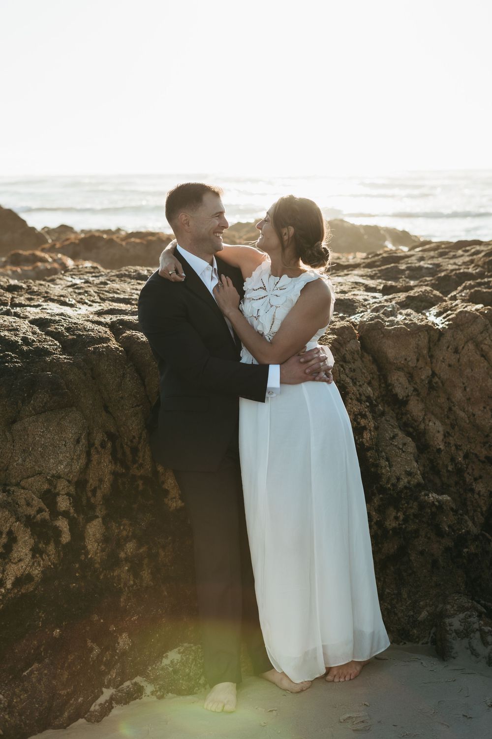 A romantic beach wedding scene with a couple embracing by rocky cliffs at sunset.