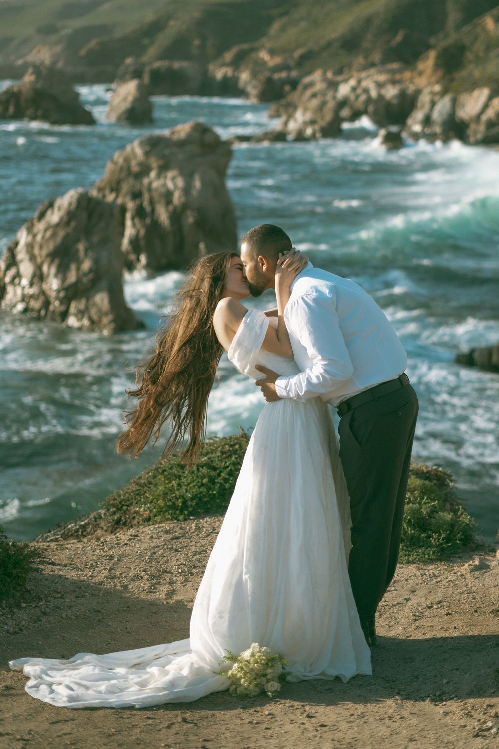 Bride and groom share a romantic kiss on a rocky coastal cliff, her white gown flowing in the ocean breeze.