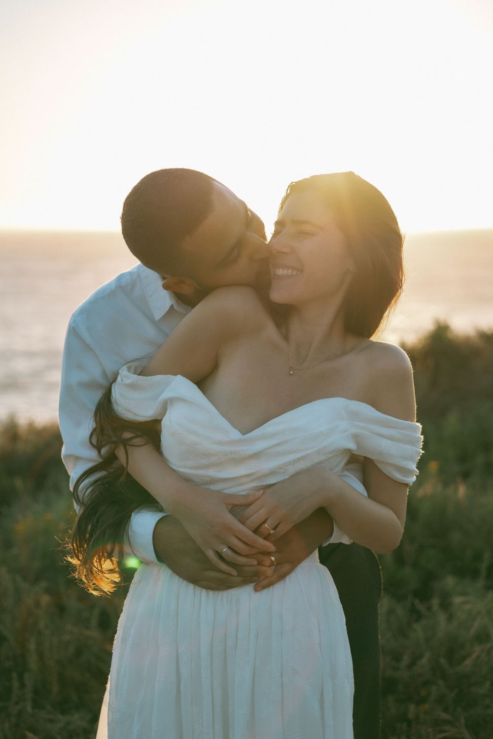 Couple embraces at sunset, groom kissing bride in off-shoulder white dress with ocean backdrop.