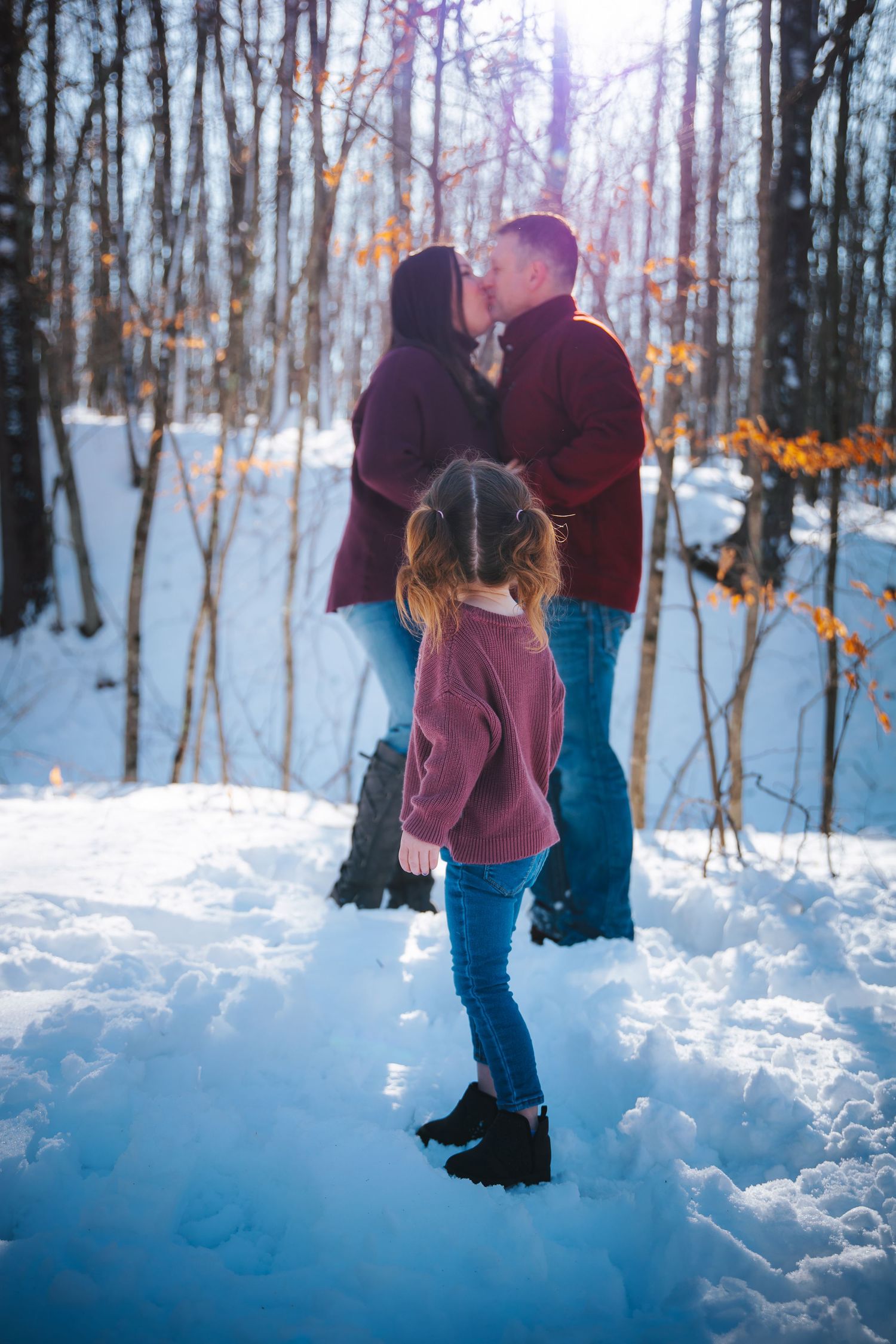 A couple shares an intimate moment in a snowy forest while a child in a burgundy sweater stands nearby.