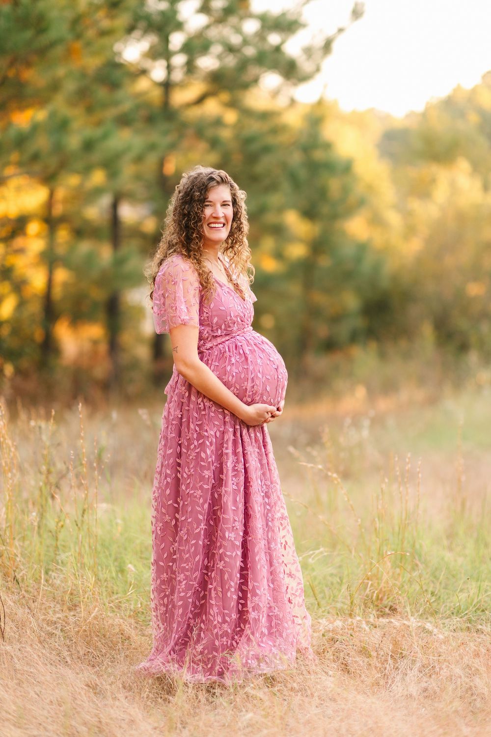 A pregnant woman in a pink lace maxi dress poses in a golden sunlit field during sunset.