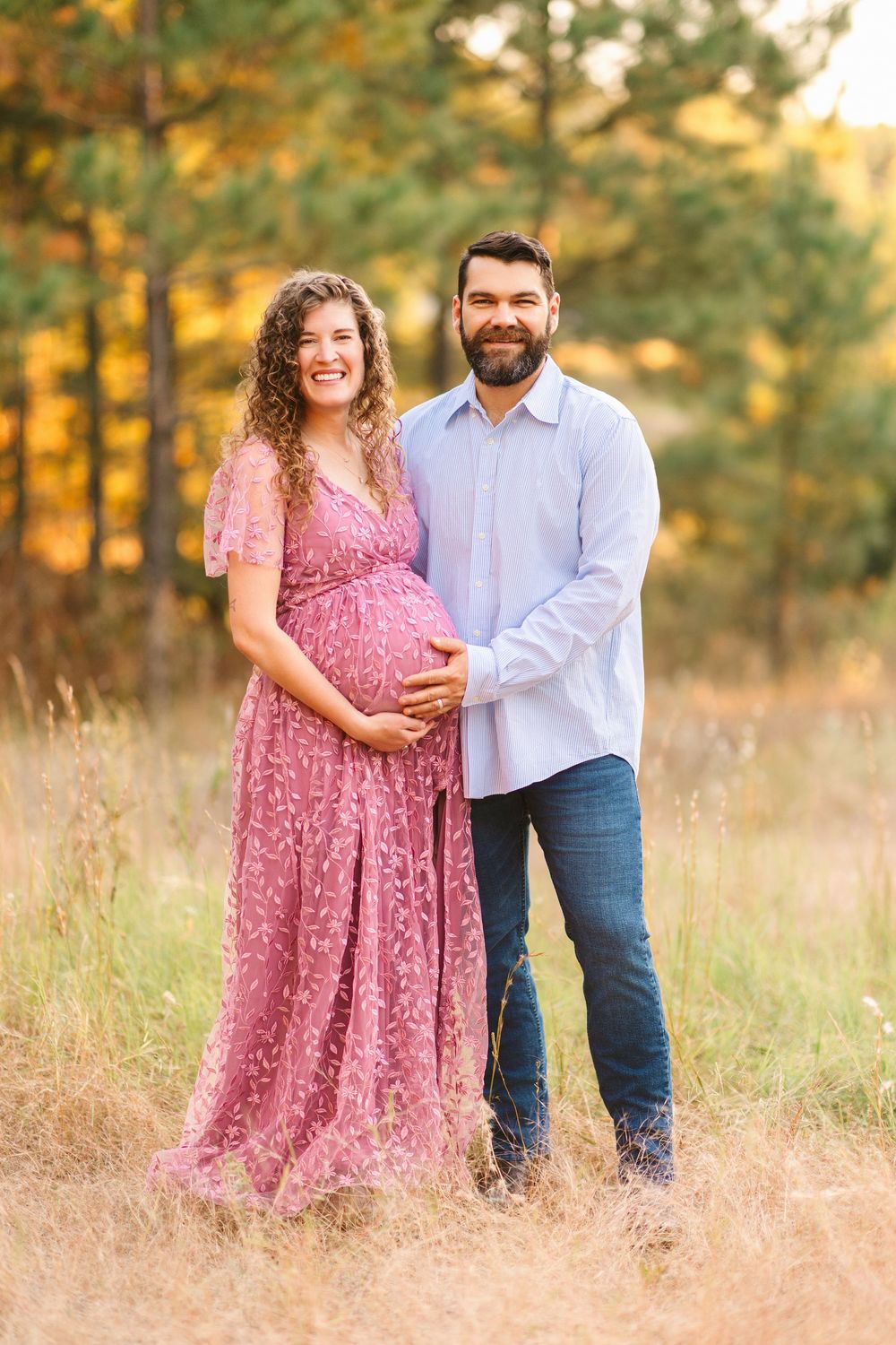 A couple embraces in a sun-lit field during golden hour while wearing pink and white clothing.