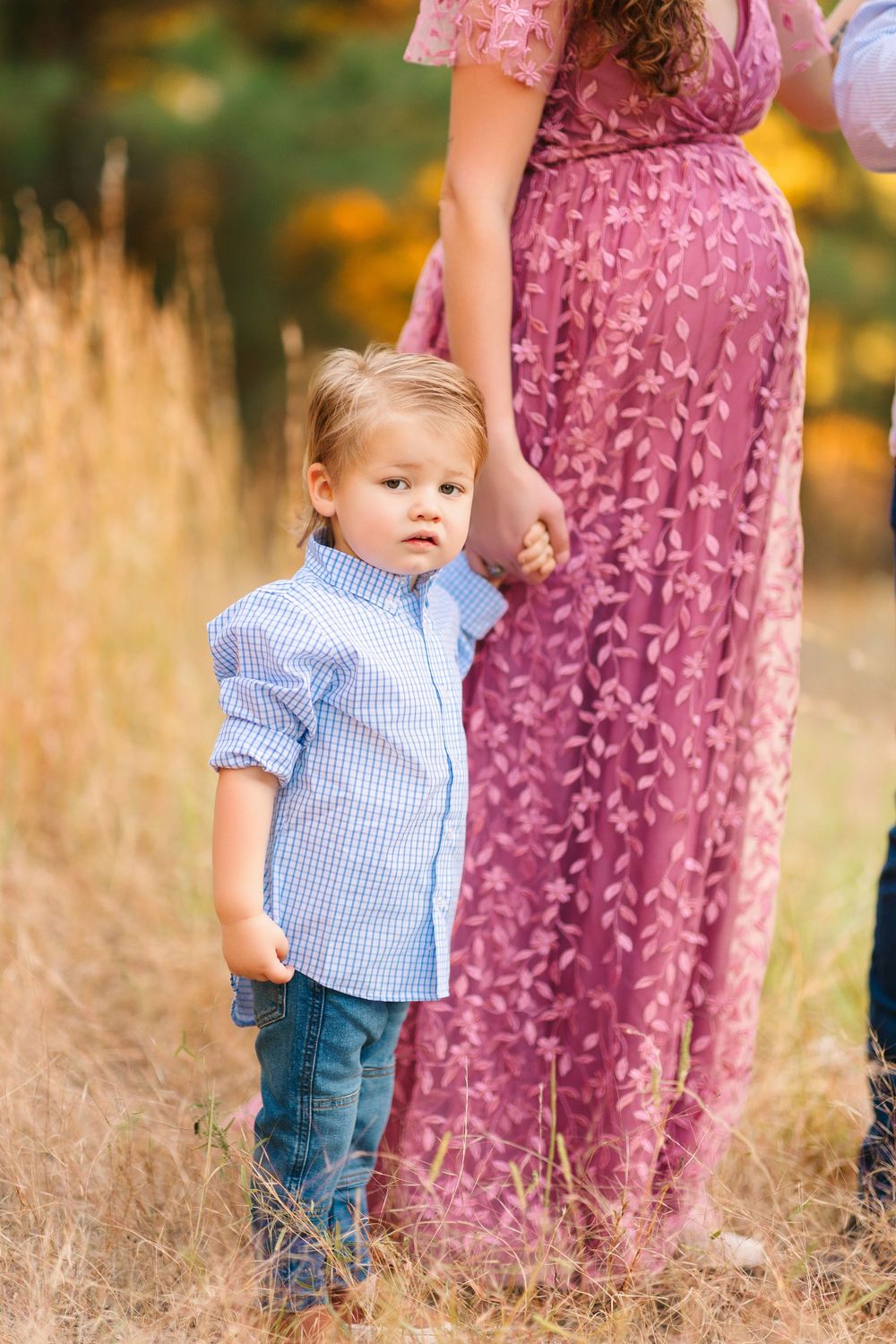 Child in blue gingham shirt and jeans stands in tall grass beside someone wearing a pink floral dress at sunset.
