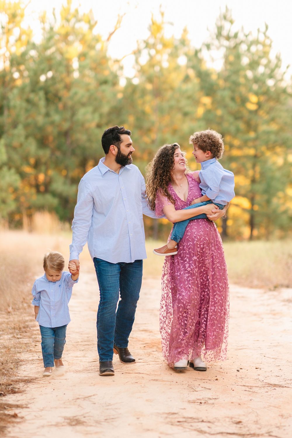 A family in coordinated outfits walks together on a dirt path surrounded by pine trees at golden hour.