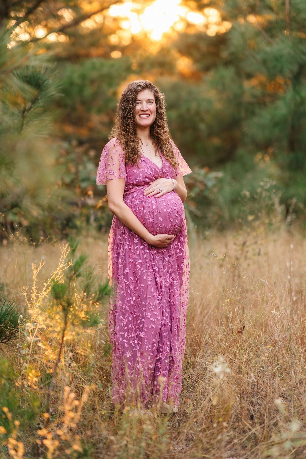 A pregnant woman in a pink floral dress stands in a golden field at sunset.