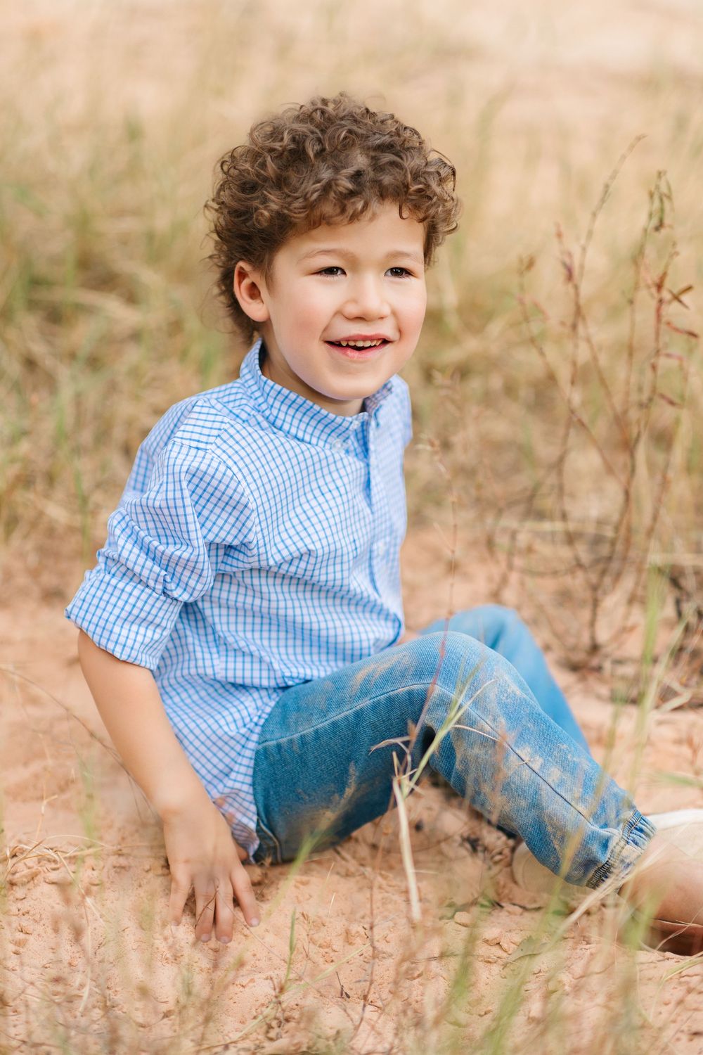 Child in blue checkered shirt and jeans playing joyfully on the ground in a sunlit field.
