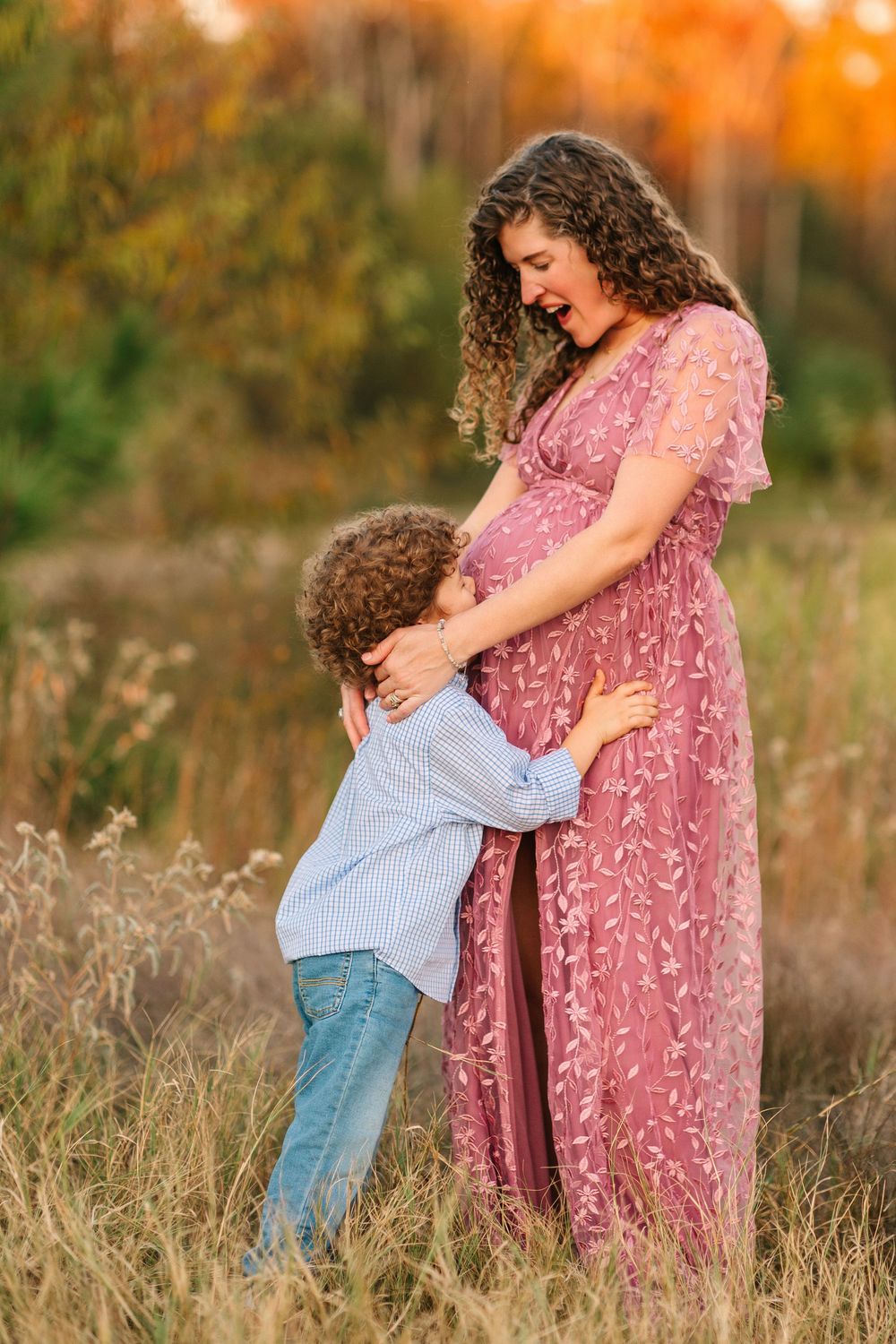 A sweet moment captured as a child hugs a pregnant belly in a grassy field at golden hour.
