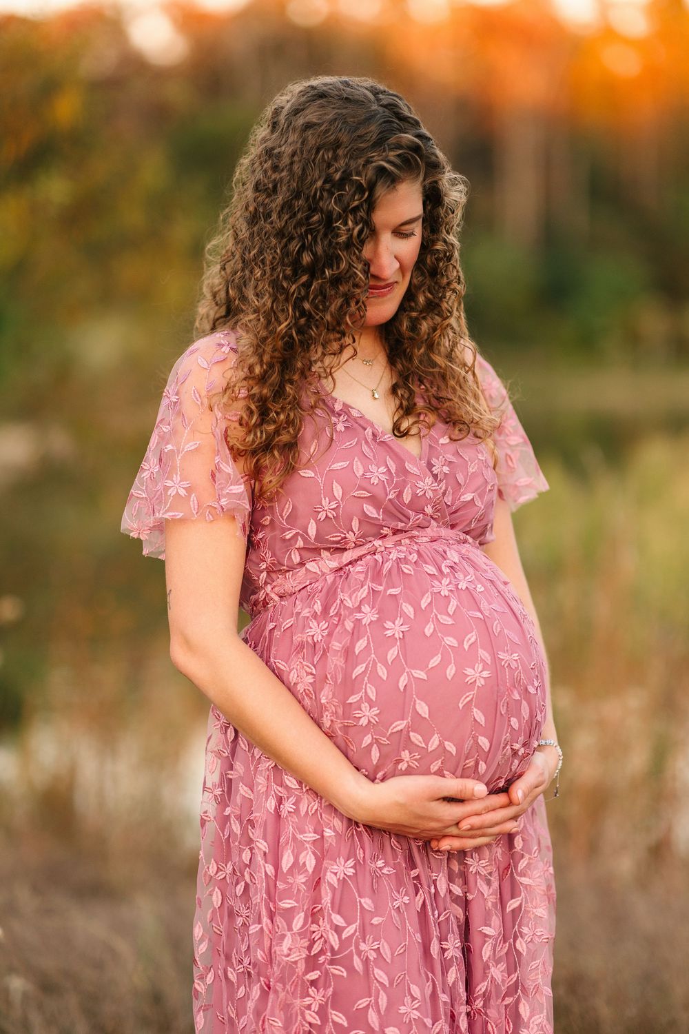 Expectant mother in pink floral dress caressing baby bump during golden hour outdoor maternity photoshoot.